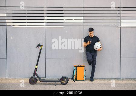 Full body front view of young delivery man with helmet using mobile phone leaning on wall near thermal backpack and electric push scooter on road Stock Photo
