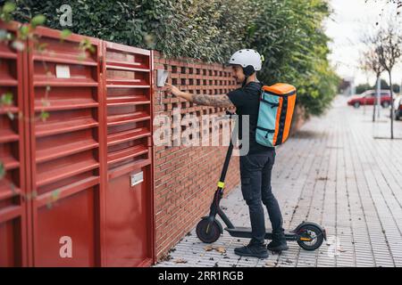 Full body side view of young delivery man in helmet with thermal backpack on electric kick scooter ringing doorbell of house Stock Photo