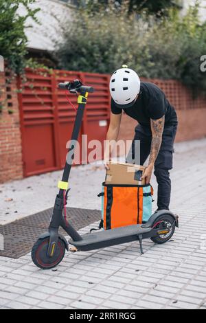 Unrecognizable delivery man in protective helmet removing package from thermal bag near electric kick scooter on pavement in town Stock Photo