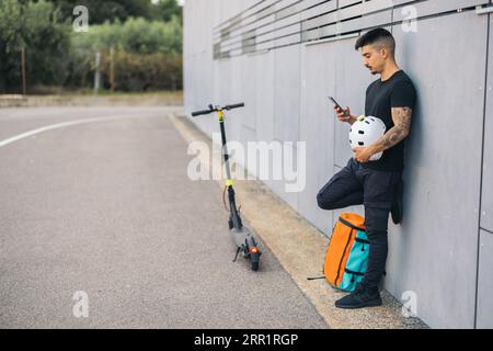 Full body side view of young delivery man with helmet using mobile phone leaning on wall near thermal backpack and electric push scooter on road Stock Photo