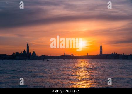 Picturesque scenery of sundown orange sky with dark clouds over cityscape with towers multistoried buildings and rippling shiny seawater with boat dur Stock Photo