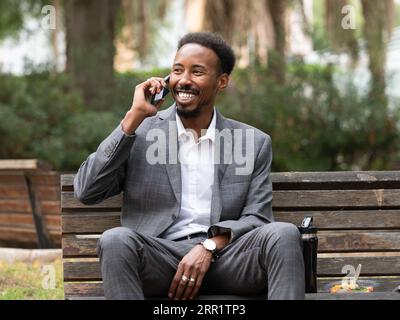 Young African American male looking away while sitting with crossed legs on bench in green park and speaking on mobile phone in daylight Stock Photo