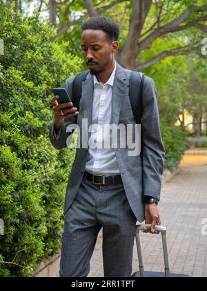 Confident young African American male entrepreneur looking at screen of mobile phone while walking on paved road with suitcase and reading messages Stock Photo