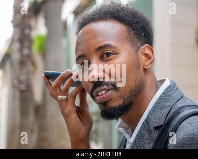 Bearded African American man in formal jacket listening to voice message on smartphone while looking away on blurred sunny street Stock Photo