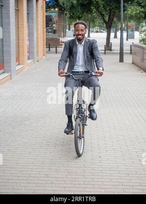 Happy male in formal suit backpack looking at camera while riding bicycle on pavement on urban street Stock Photo