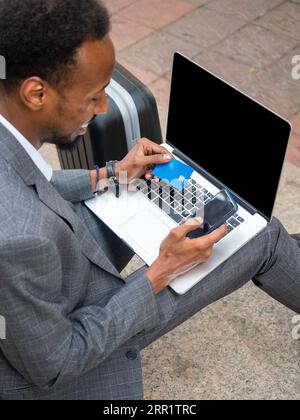 High angle of satisfied African American male in formal wear using cellphone for paying with plastic card while sitting on street with laptop Stock Photo