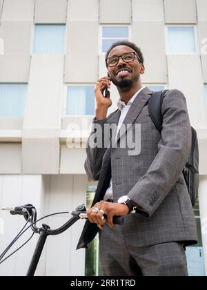 From below of African American male in formal suit standing with bicycle on city street and talking on mobile phone Stock Photo