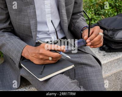From above of anonymous black man in formal wear sitting on bench on street and holding eyeglasses and pen with notebook Stock Photo