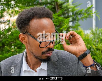 Attentive black male in stylish jacket touching glasses while looking away near green tree on street on sunny day Stock Photo
