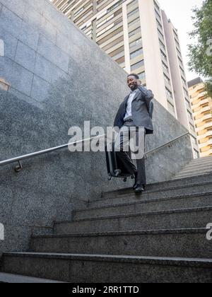 From below full body of happy African American male in formal clothes speaking on mobile phone while walking down the stairs with suitcase and backpac Stock Photo