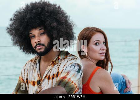 Confident multiracial couple in casual clothes looking at camera while sitting near sea on sunny summer day Stock Photo