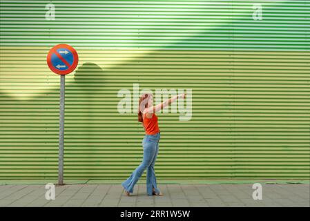 Full body side view of young female in casual clothes pointing away while standing near no parking sign and green striped wall Stock Photo