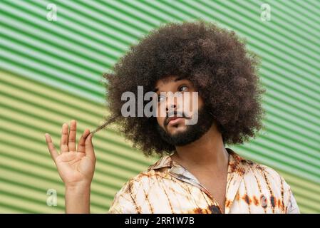 Hispanic bearded male with Afro hairstyle in Hawaiian shirt looking away  thoughtfully while touching hair against ribbed green backdrop stock photo