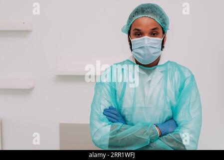 Serious ethnic bearded male doctor in medical mask robe and gloves standing near white wall with crossed arms and looking at camera Stock Photo