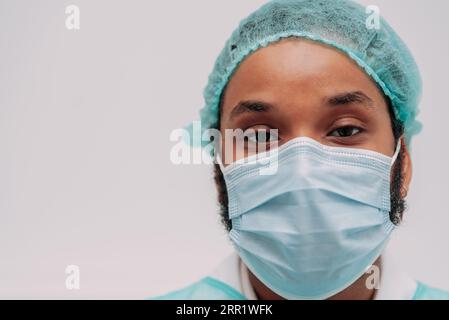 Serious ethnic bearded male doctor in medical mask robe standing near white wall looking at camera Stock Photo