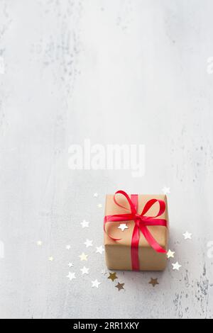 A small gift wrapped in kraft paper with a red ribbon on a light blue background, surrounded by a shiny confetti star, copy space. Christmas Stock Photo