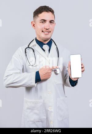 Cheerful doctor holding and pointing at cell phone screen. Young doctor showing and pointing at smatphone screen isolated Stock Photo