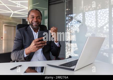 Portrait of successful winner at workplace inside office, African American man smiling and looking at camera, businessman holding phone, received online notification of winning message. Stock Photo