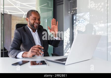 Joyful successful african american man inside office at workplace talking remotely on video call, man smiling and looking at laptop screen, waving hand in greeting gesture. Stock Photo