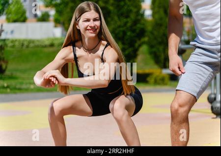 Young sporty man and woman doing workout and squatting together on an open air workout ground. Stock Photo