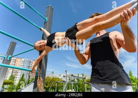 Girl doing human flag exercise with her trainer on a climbing frame. Stock Photo