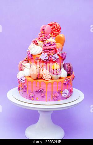 Pink, purple and orange gradient cake with cream cheese roses, macarons, cupcakes, meringues on purple plain background Stock Photo