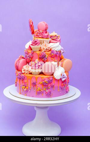 Pink, purple and orange gradient cake with cream cheese roses, macarons, cupcakes, meringues on purple plain background Stock Photo