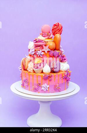 Pink, purple and orange gradient cake with cream cheese roses, macarons, cupcakes, meringues on purple plain background Stock Photo
