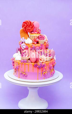 Pink, purple and orange gradient cake with cream cheese roses, macarons, cupcakes, meringues on purple plain background Stock Photo