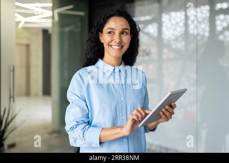 Portrait of young beautiful business woman inside office with tablet computer in hands, Hispanic smiling and looking out the window, thinking and dreaming about new projects and achievement results. Stock Photo