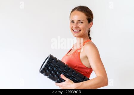 Female athlete wearing sportswear stands in front of white background holding foam roller in hand and smiling. Stock Photo