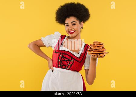 african american oktoberfest waitress in traditional costume holding pretzels and smiling on yellow Stock Photo