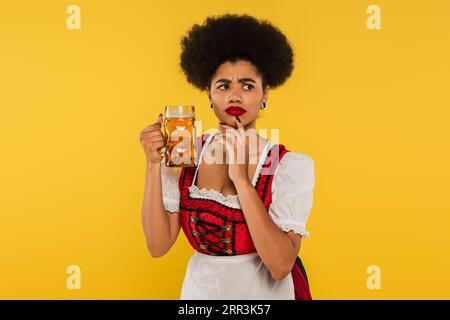 thoughtful african american oktoberfest waitress in dirndl standing with beer mug on yellow Stock Photo