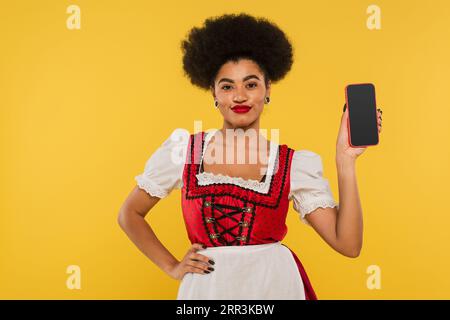 african american oktoberfest waitress in dirndl showing smartphone with blank screen on yellow Stock Photo