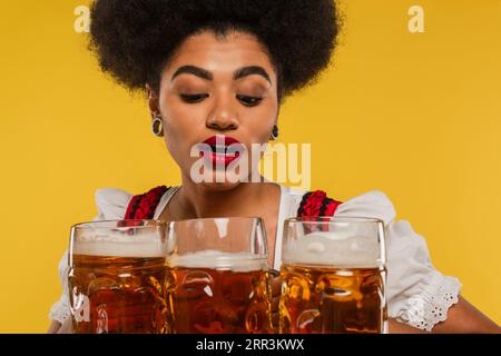 pretty african american oktoberfest waitress in bavarian attire looking at full beer mugs on yellow Stock Photo