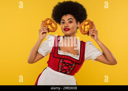 smiling african american oktoberfest waitress in dirndl showing delicious pretzels on yellow Stock Photo