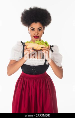 african american oktoberfest waitress in bavarian costume holding hot dog and smiling on white Stock Photo
