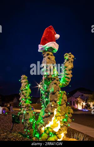 A totem pole cactus lit up with Christmas lights and a Santa hat in front of a home in the evening. Stock Photo