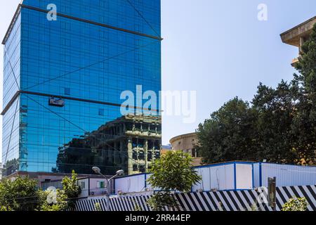 The blue glass façade of a high-rise in Tbilisi, Georgia, reflects the columns and storeys of a neighbouring building. A work gondola hangs on the façade. On the right, a round building with a cornice stands behind trees, and a concrete building cantilevers out above it. Site containers stand behind a black-and-white striped construction fence in the foreground. Stock Photo