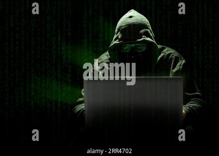 A hooded hacker committing a cyber attack with a laptop, against a background of matrix-style computer code. Concept of computer crimes in the digital Stock Photo