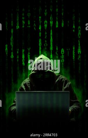 A hooded hacker works on a cyber attack with a laptop, against a background of matrix style computer code where a skull can be seen. Cybercrimes. Stock Photo