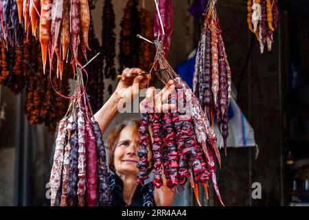 A vendor cuts strands of churchkhela (Georgian ჩურჩხელა) at her stall at the Dezerter Bazaar (Deserters' Market) in Tbilisi, Georgia. With raised arms she holds a bundle and a pair of scissors and smiles. The strands in red, purple, orange and white hang in dense clusters on strings. Churchkhela is a Georgian confection. The nuts are coated in pelamushi, a glaze of boiled-down grape juice with starch flour and no sugar. To make it, walnuts or hazelnuts are threaded onto a string, dipped repeatedly into pelamushi and then dried. Churchkhela is a classic provision for hiking in the Caucasus. The Stock Photo