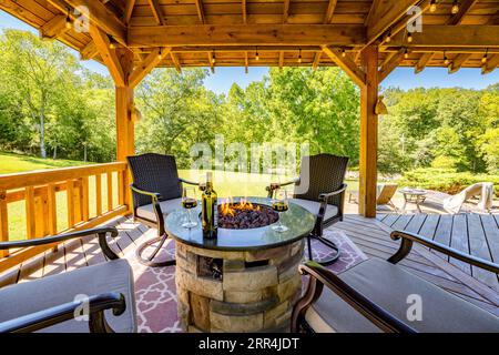 An outdoor living scene with four chairs surrounding a firepit under a pavilion at a country home. A wine bottle & stemmed glasses sit around fire. Stock Photo