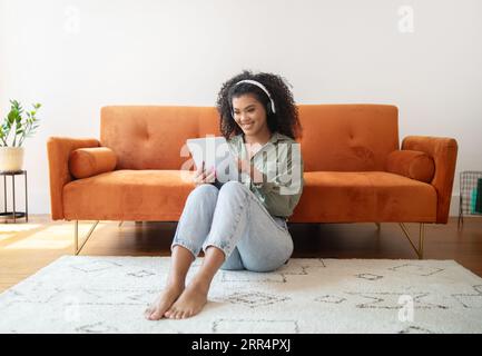 Black Young Woman Using Digital Tablet Wearing Wireless Headphones Indoors Stock Photo