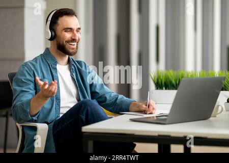 Smiling Businessman In Headphones Making Video Call And Taking Notes At Workplace Stock Photo