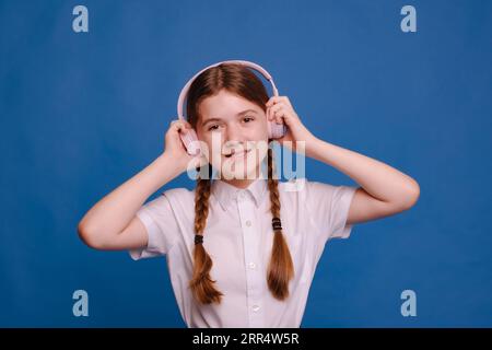 happy school girl with hairstyle two braids listen online lessons with headphones on blue background Stock Photo