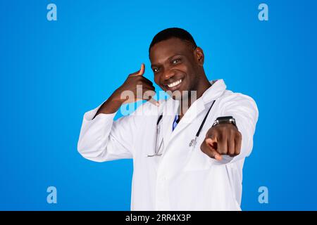 Cheerful young african american man doctor in white coat pointing finger at camera and sign call me with hand Stock Photo