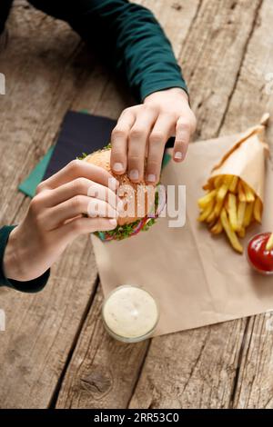 Burger in hands, french fries, sauces, spices on a wooden background. Fast food concept. Stock Photo