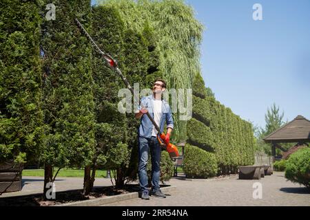 Male gardener in protective glasses using hand electric trimmer for cutting trees in summer. Low angle view of strong worker in denim clothes shaping thujas on backyard. Concept of seasonal work. Stock Photo