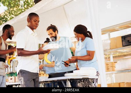 Diverse volunteers collaborate at homeless center, distributing essentials, serving free food to underprivileged, supporting hunger relief. Group of voluntary people provide aid to the poor and needy. Stock Photo